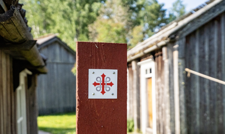 Logo föreställande ett kors på en röd stolpe. I bakgrunden gamla hus av grånat trävirke.