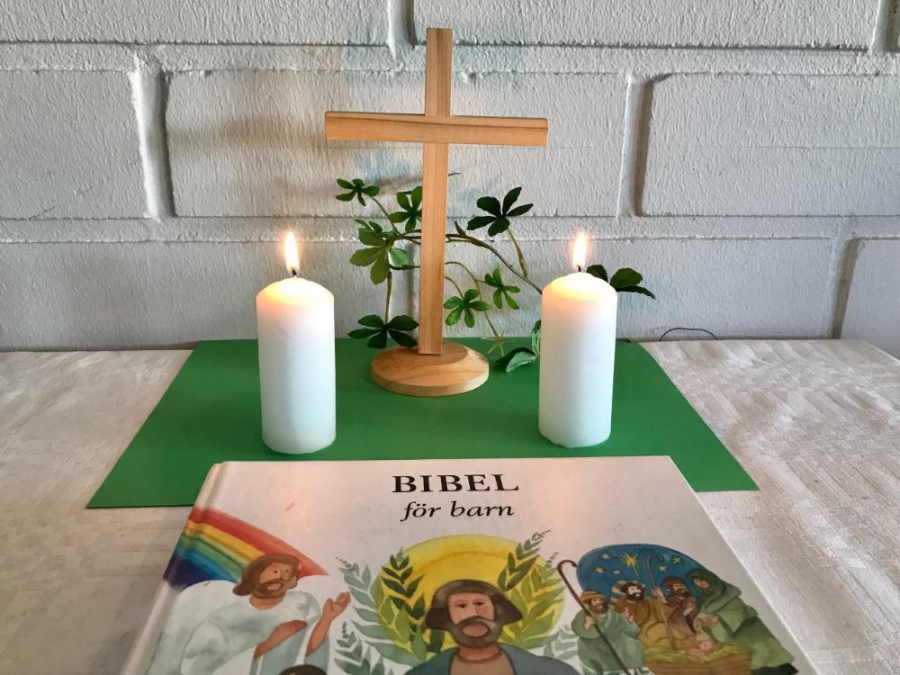 Altarbord med ett träkors och Bibel för barn. Två vita ljus brinner.