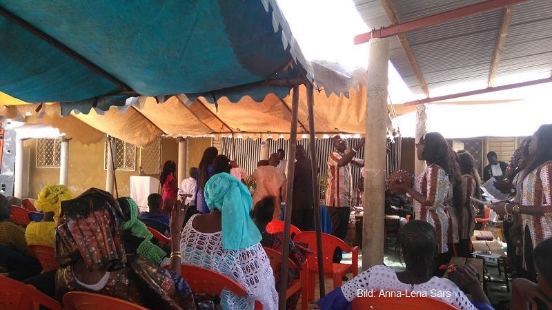 Gudstjänst i ett tält i Senegal. Människor som sitter. Kör som står och sjunger. Människor på altaret.
