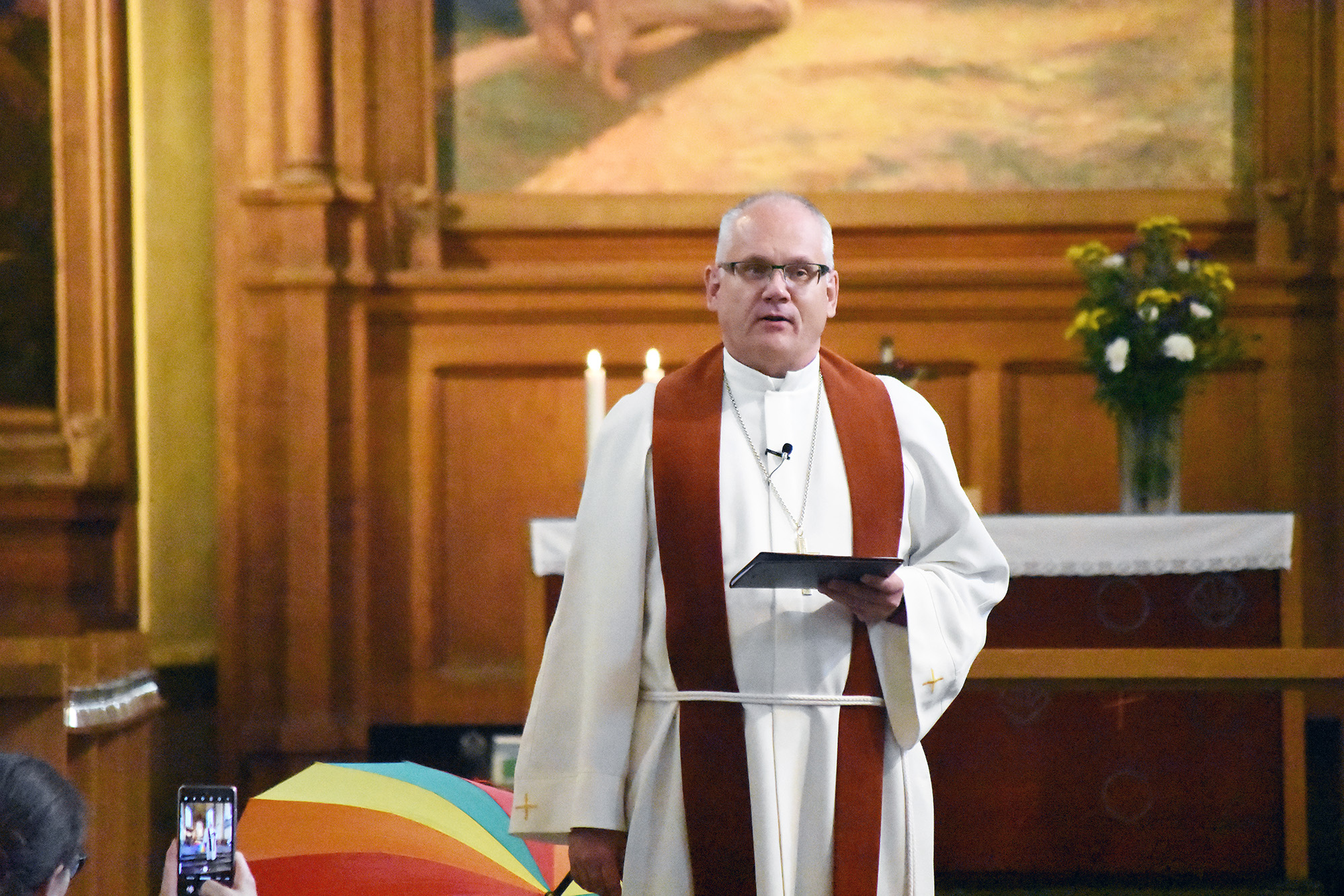 Man iklädd alba pratar i en kyrka. I bakgrunden syns ett regnbågsfärgat paraply.