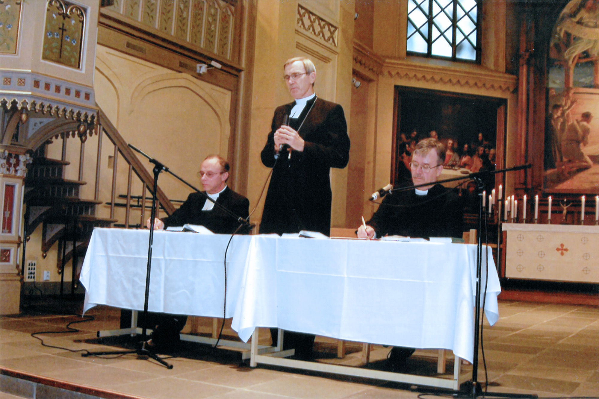 Tre män i prästkläder sitter vid ett bord framme i en kyrka.