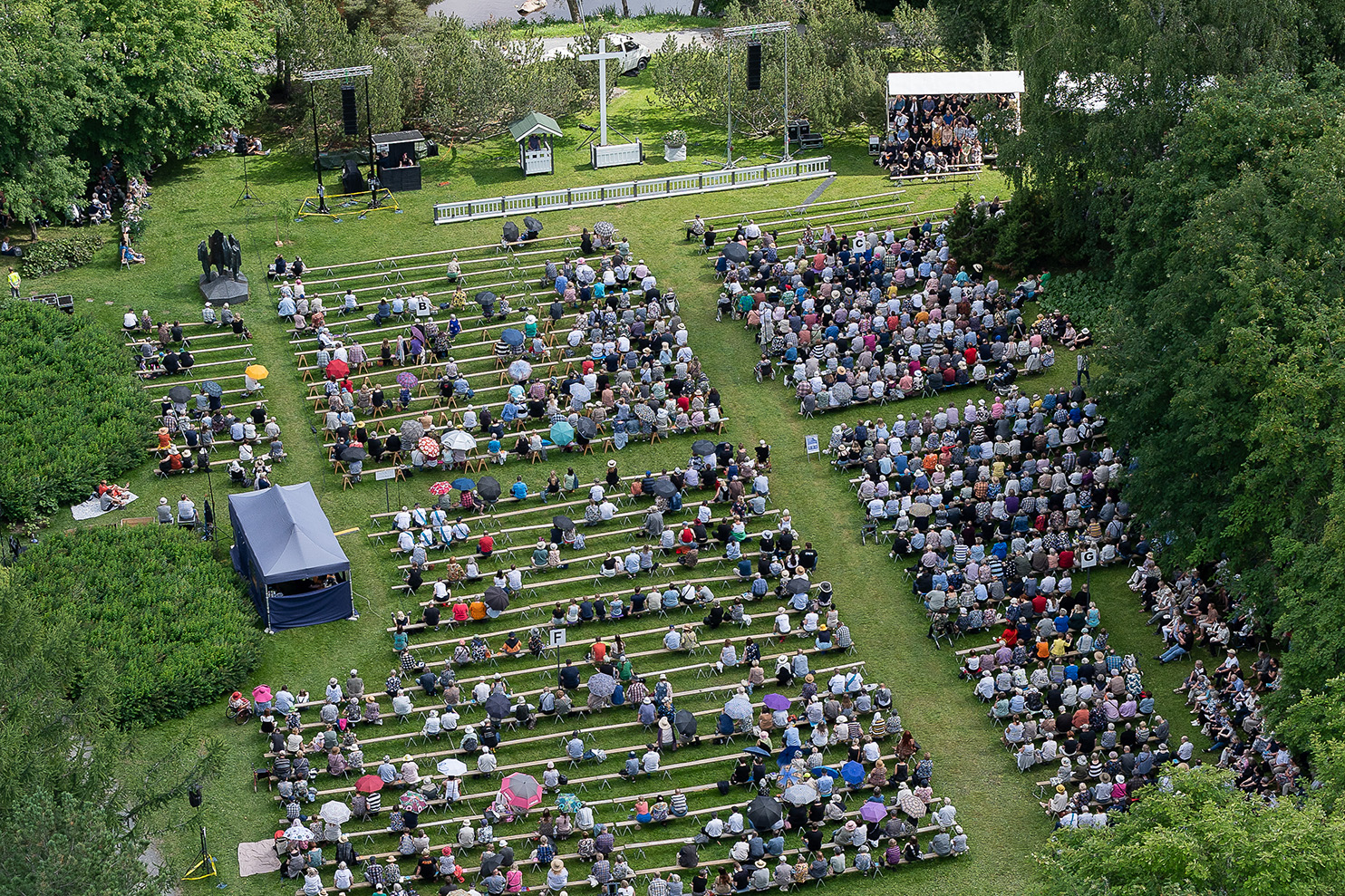 Massor av människor sitter på bänkar på en stor grön gräsmatta.