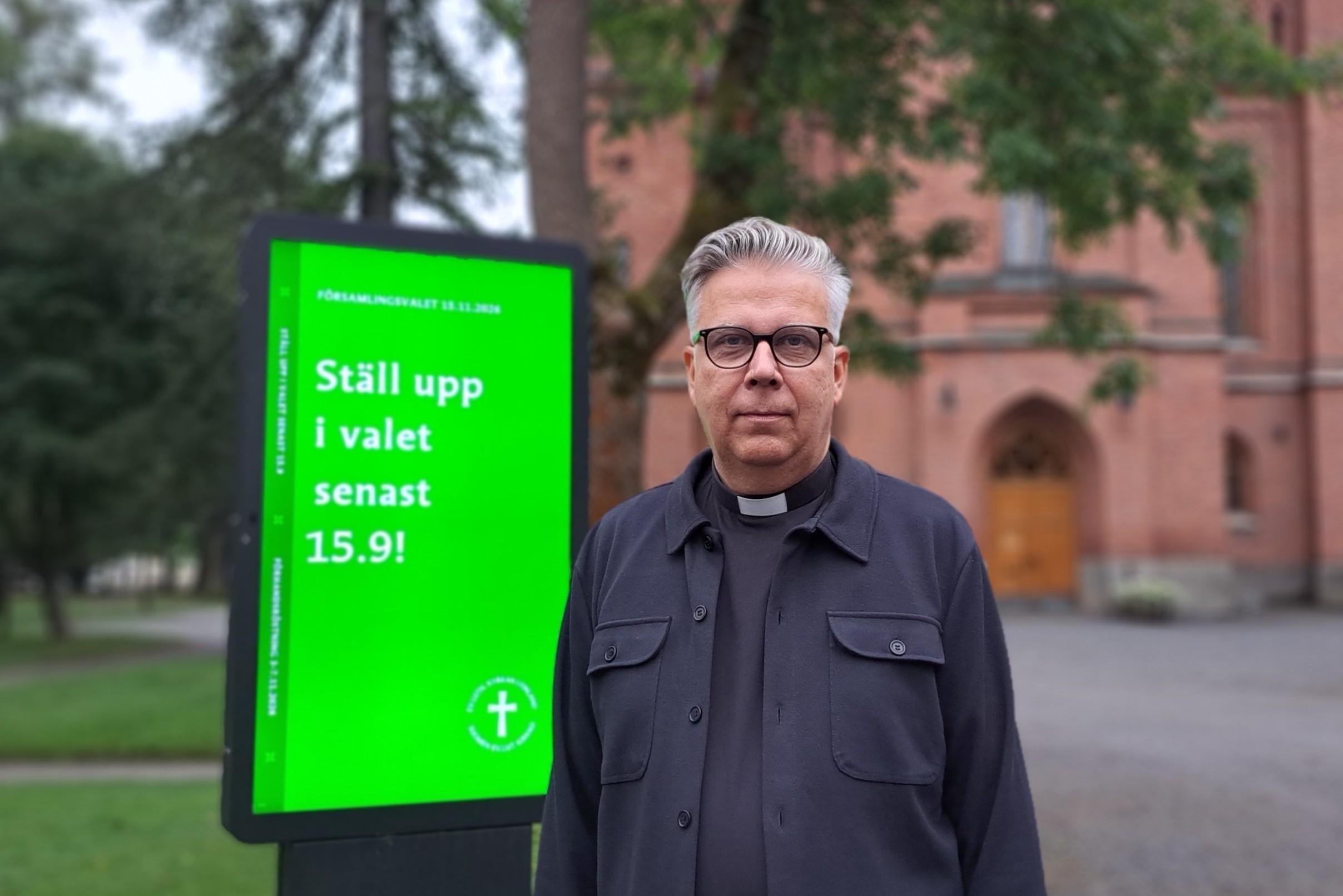 Man står bredvid en ljustavla med texten Ställ upp i valet senast 15.9. I bakgrunden en röd tegelkyrka.