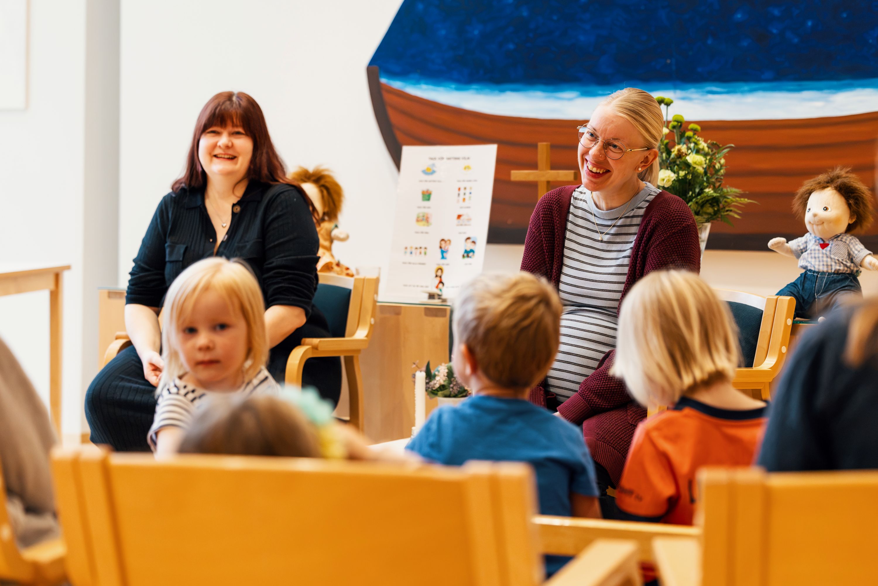 Barn och vuxna sitter samlade i en ring i ett ljust församlingsutrymme. I bakgrunden syns ett kors, blommor...