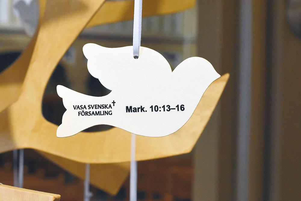 Vit duva av trä som hänger på en gren. Text: Vasa svenska församling. Mark. 10:13-16.