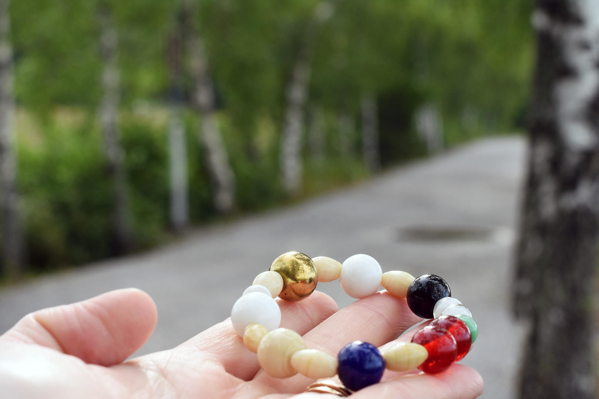 Hand som håller i ett armband med olikfärgade pärlor. I bakgunden en väg och skog.