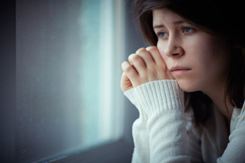 En kvinna som ser ledsen ut tittar ut genom ett fönster. Länken leder till webbsidan om sorg och död.