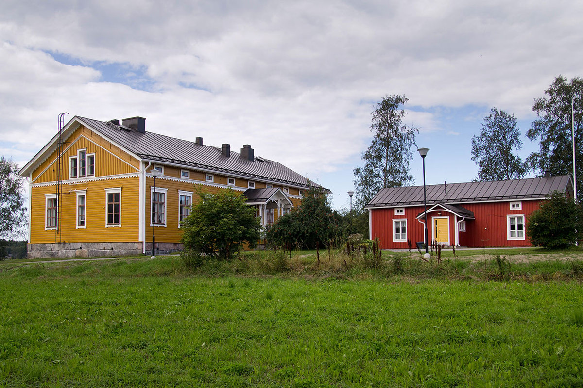 Gul avlång prästgård av trä med brunt plåttak. På samma gårdsplan syns en mindre röd träbyggnad.