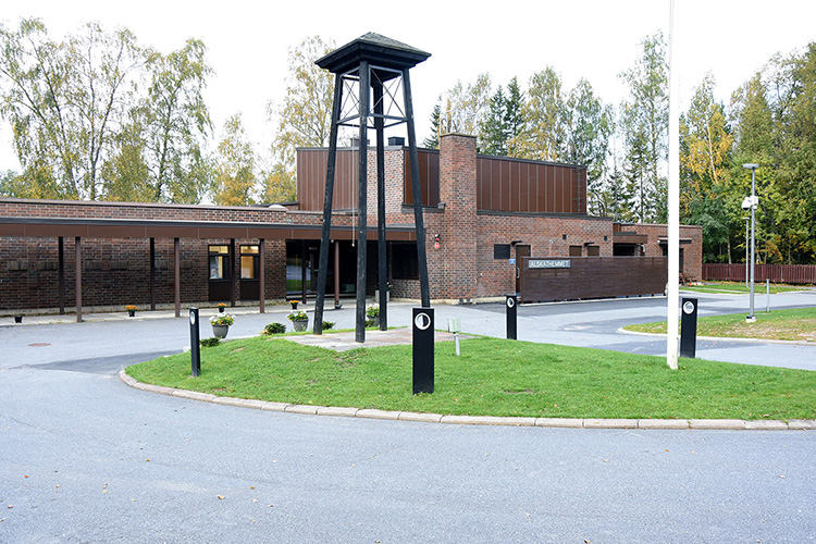 Mörkbrun tegelbyggnad. På gårdsplanen framför finns en hög klockstapel.