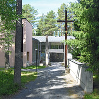 Gerby församlingshem Ingång till fastighet som skymtar bakom en träddunge. På gårdsplanen finns ett stort kors.