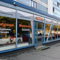 Cross Låg vit tegelbyggnad med stora fönster framtill tejpade med texten missionsshop och Cross
