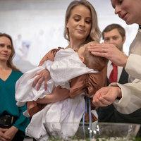 Kvinna håller ett barn i famnen medan prästen sätter vatten på huvudet. Länken leder till kyrkans webbsida om dop. 