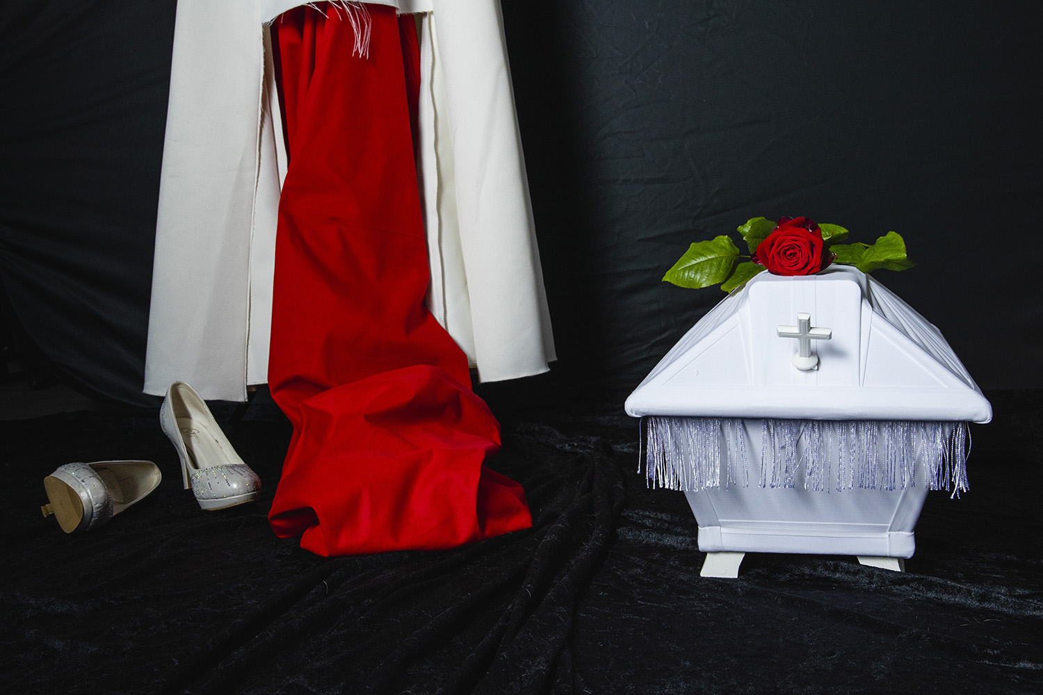 En vit barnkista med röd ros på golvet. I bakgrunden syns en trasig brudklänning.