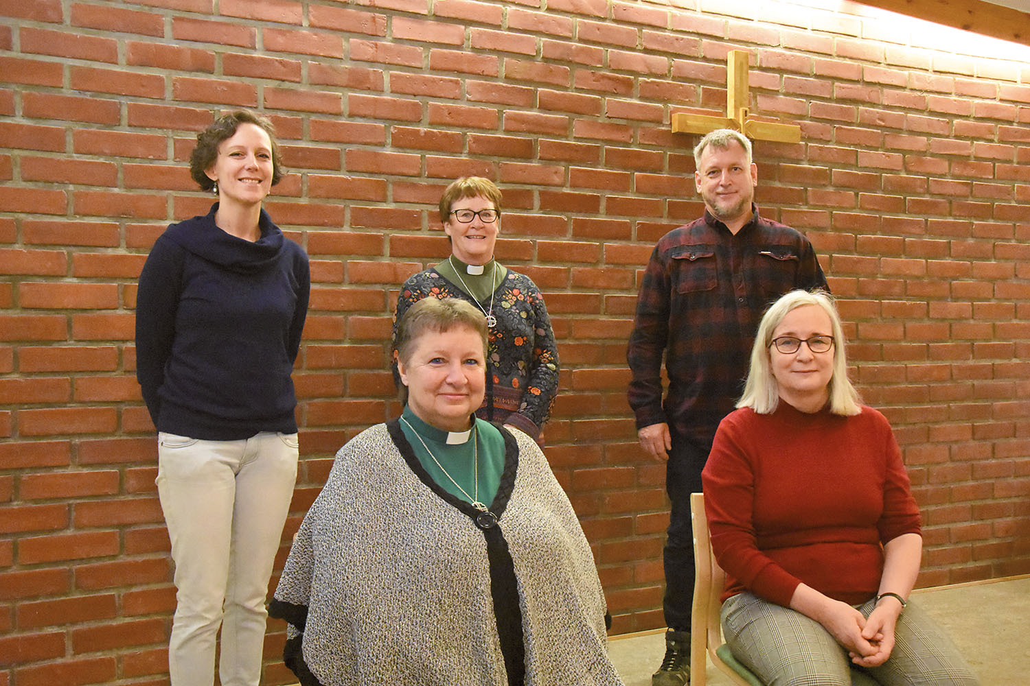 Fyra kvinnor och en man står i en gles klunga. I bakgrunden finns ett träkors på en tegelvägg.