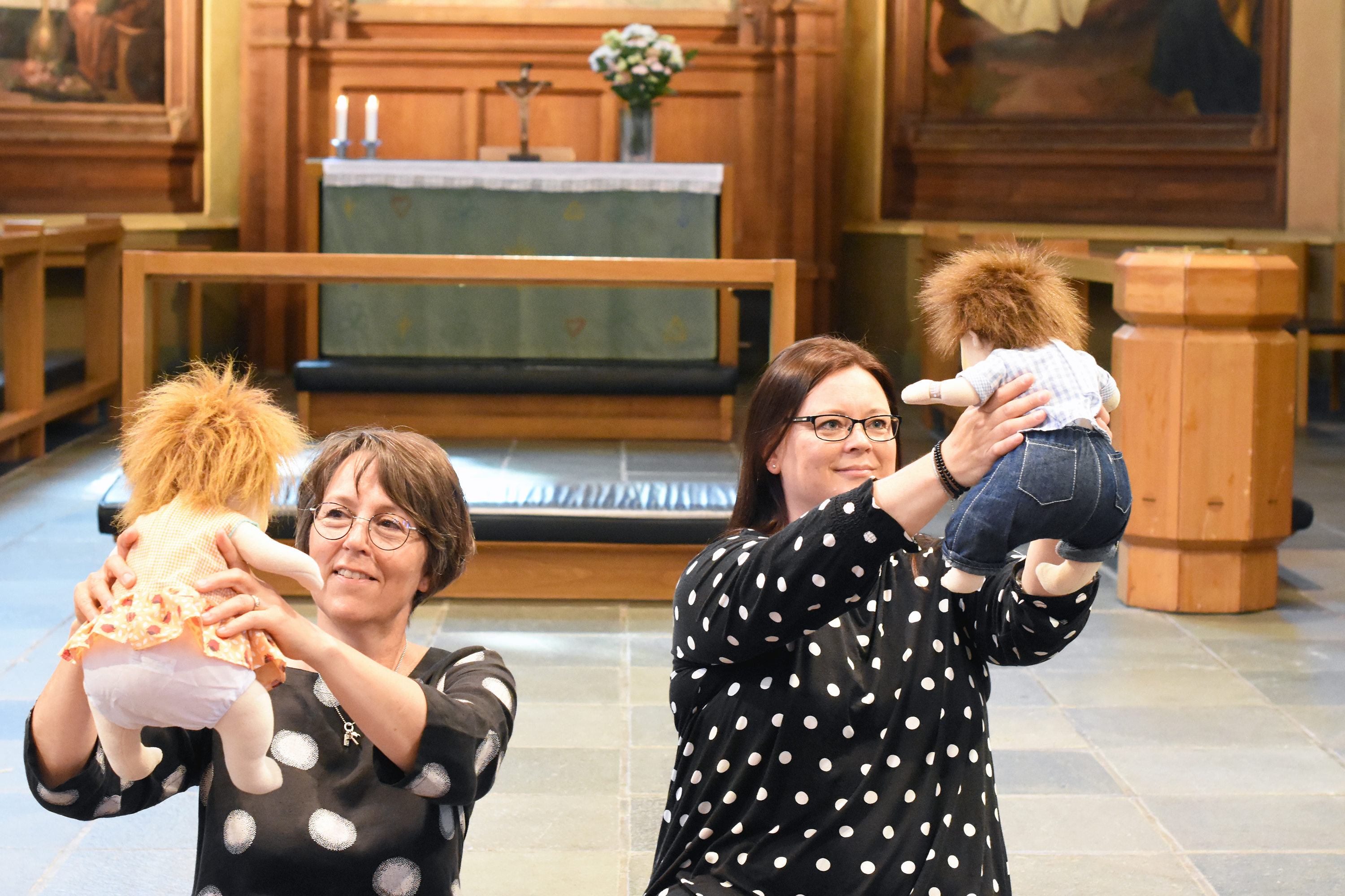 Två kvinnor sitter i en kyrka med mjuka dockor i händerna höjda ovanför huvudena. De skrattar och ser glada...