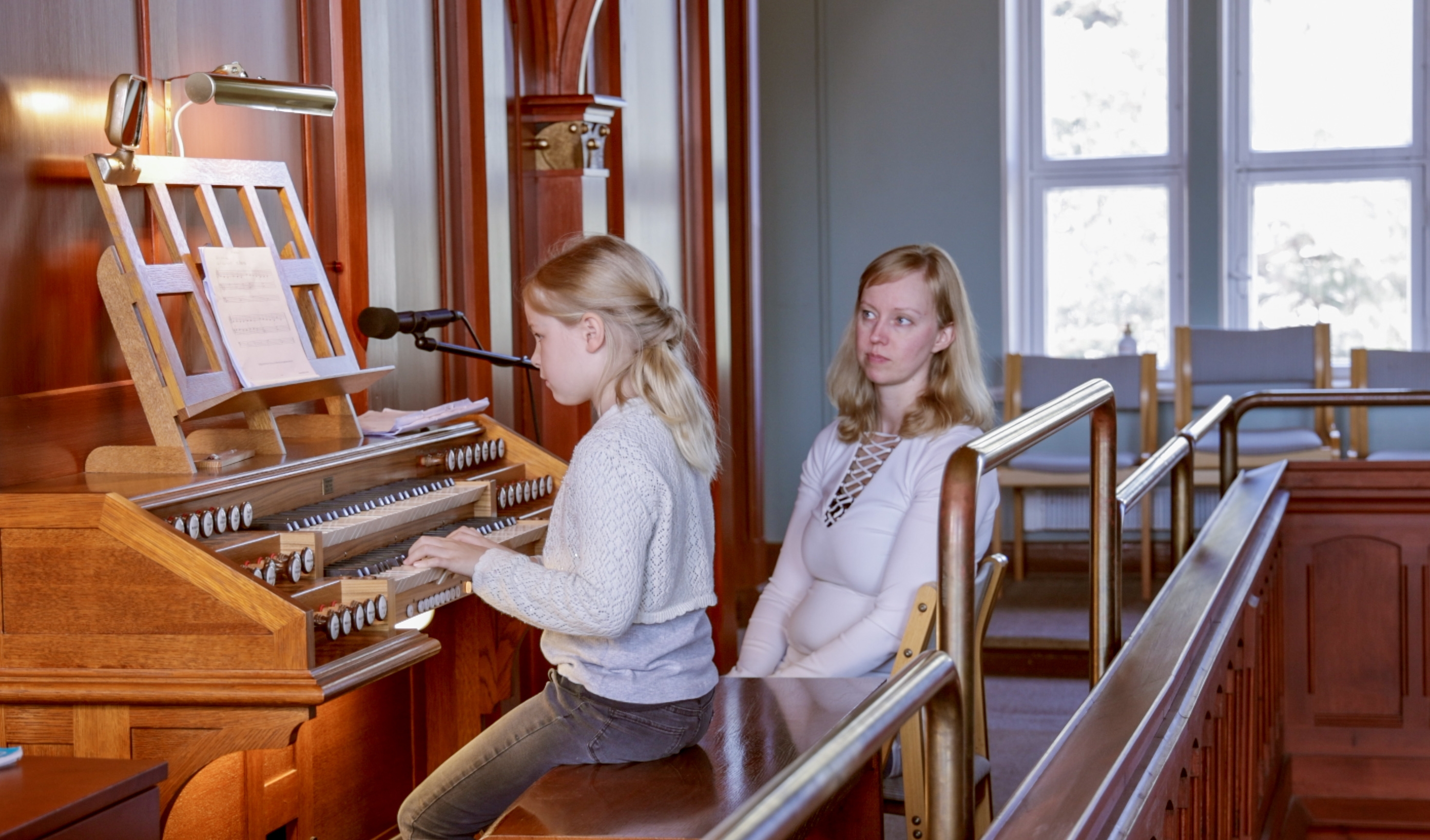 Flicka spelar orgel. Kvinna sitter bredvid på en stol.