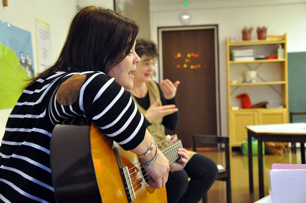 Två kvinnor som sjunger. En spelar gitarr. De är i ett rum med leksaker i.
