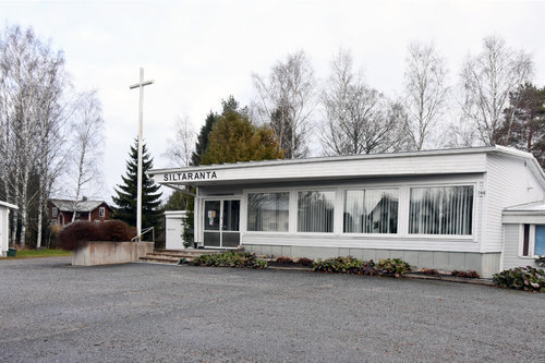 Låg, vit träbyggnad med stora fönster. På gårdsplanen finns ett stort vitt kors.