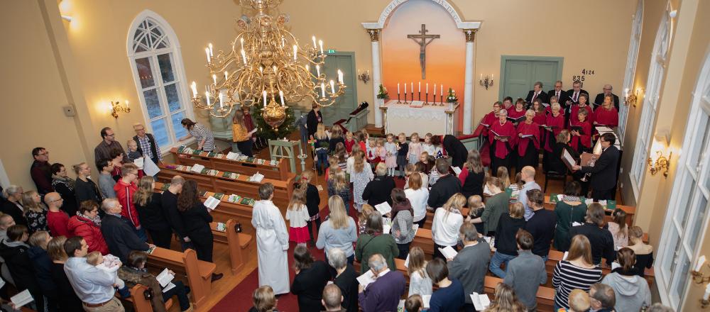 Massor med människor står och rör sig inne i en kyrka. Framme i kyrkan sjunger en vuxenkör och en barnkör.