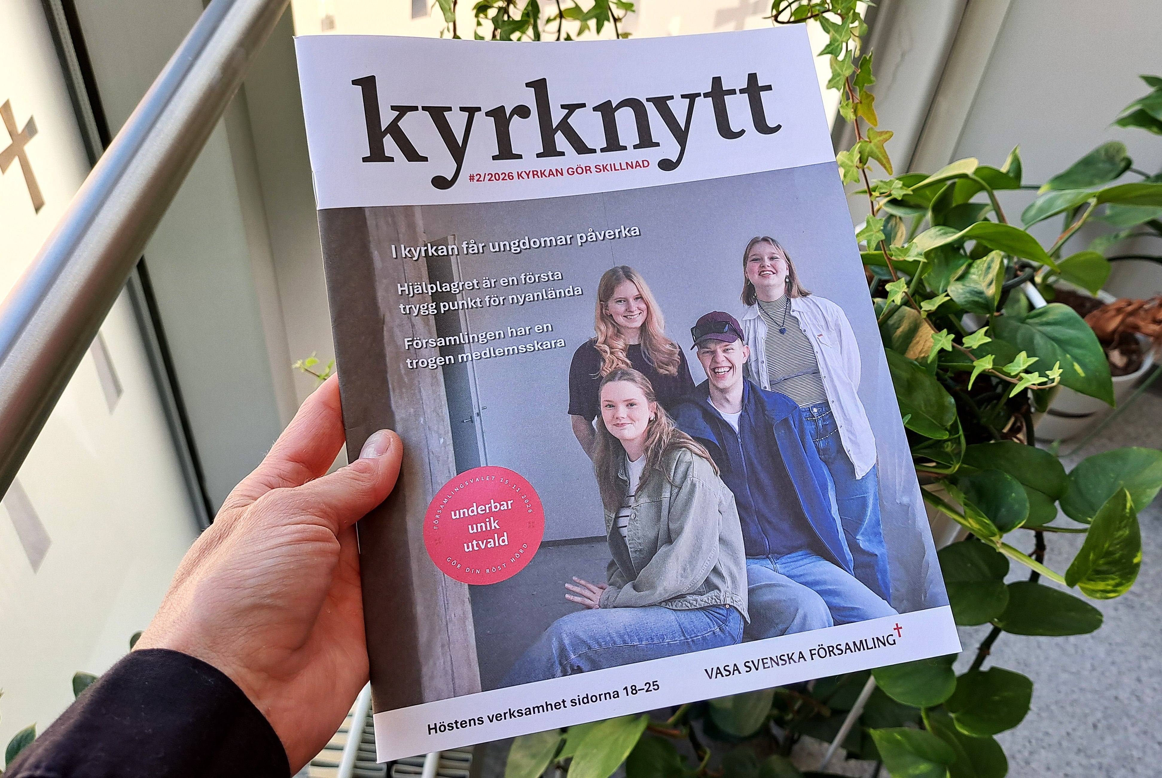 Hand som håller i tidningen Kyrknytt. I bakgrunden grönväxter.
