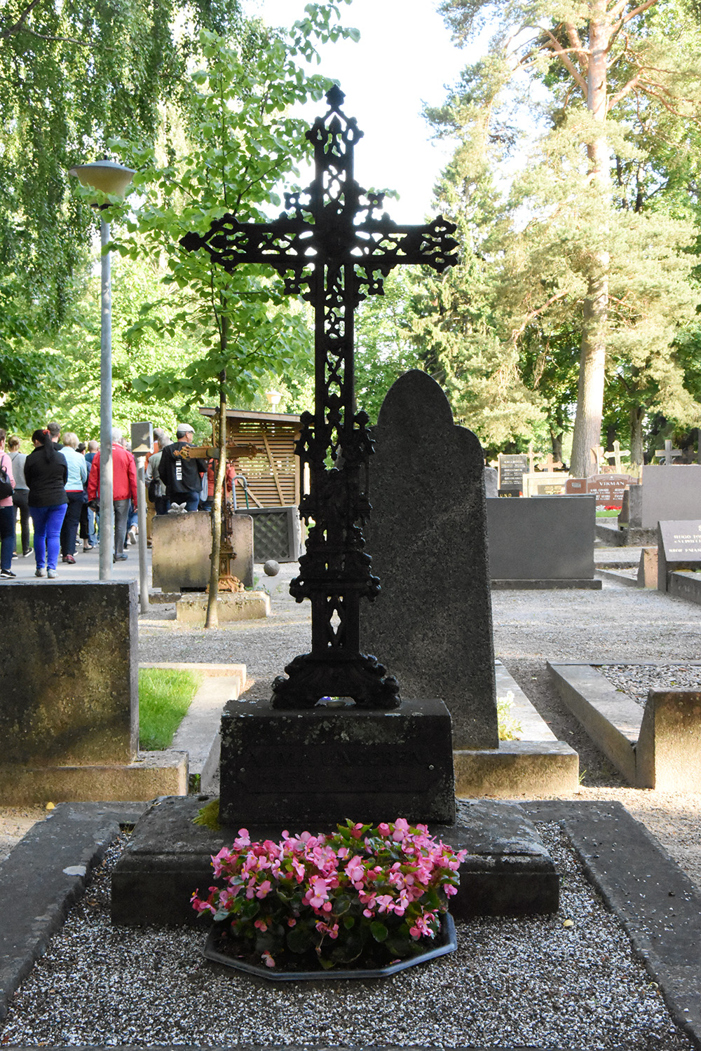 Svart utsmyckat tunt kors av järn på en begravninsplats. Framför korset finns rosa blommor.