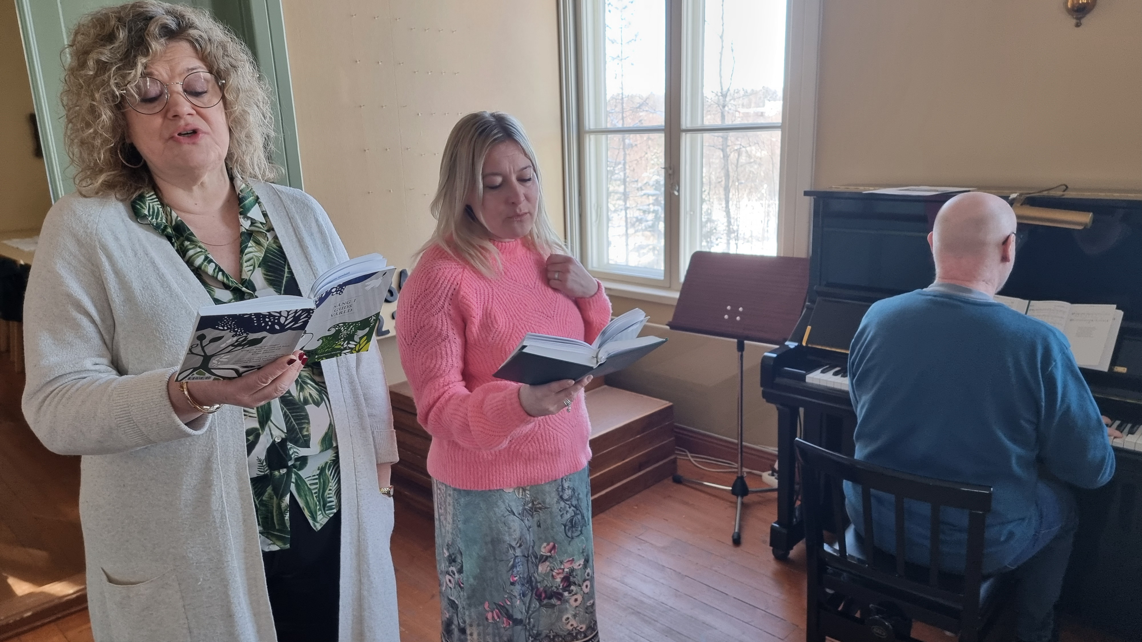 Två kvinnor sjunger i en kyrka och en man spelar piano.