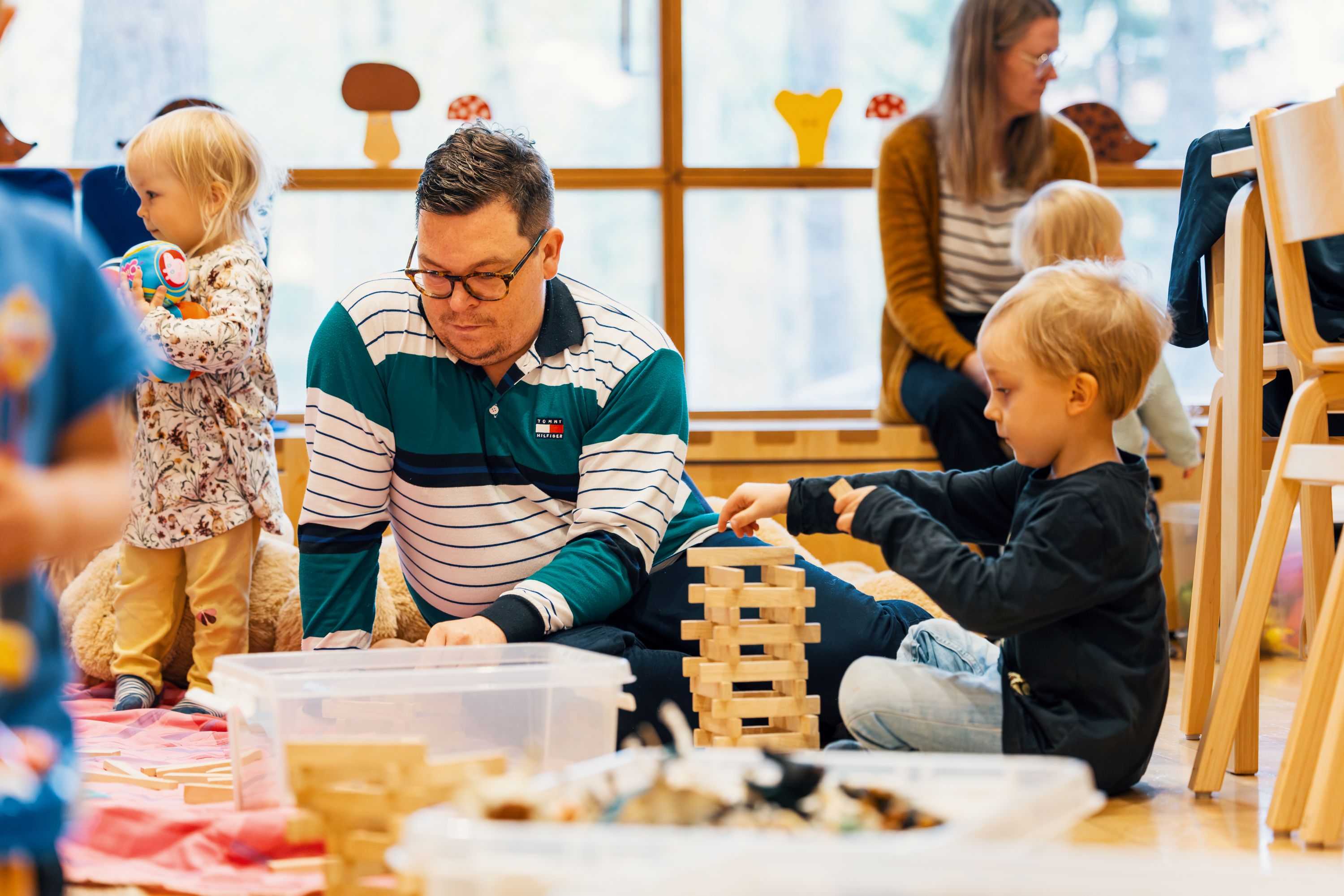 Barn och vuxna sitter på golvet och leker med byggklossar och andra leksaker i ett ljust rum. Ett barn bygger ett torn av träklossar medan övriga deltagare är sysselsatta med lek i bakgrunden.