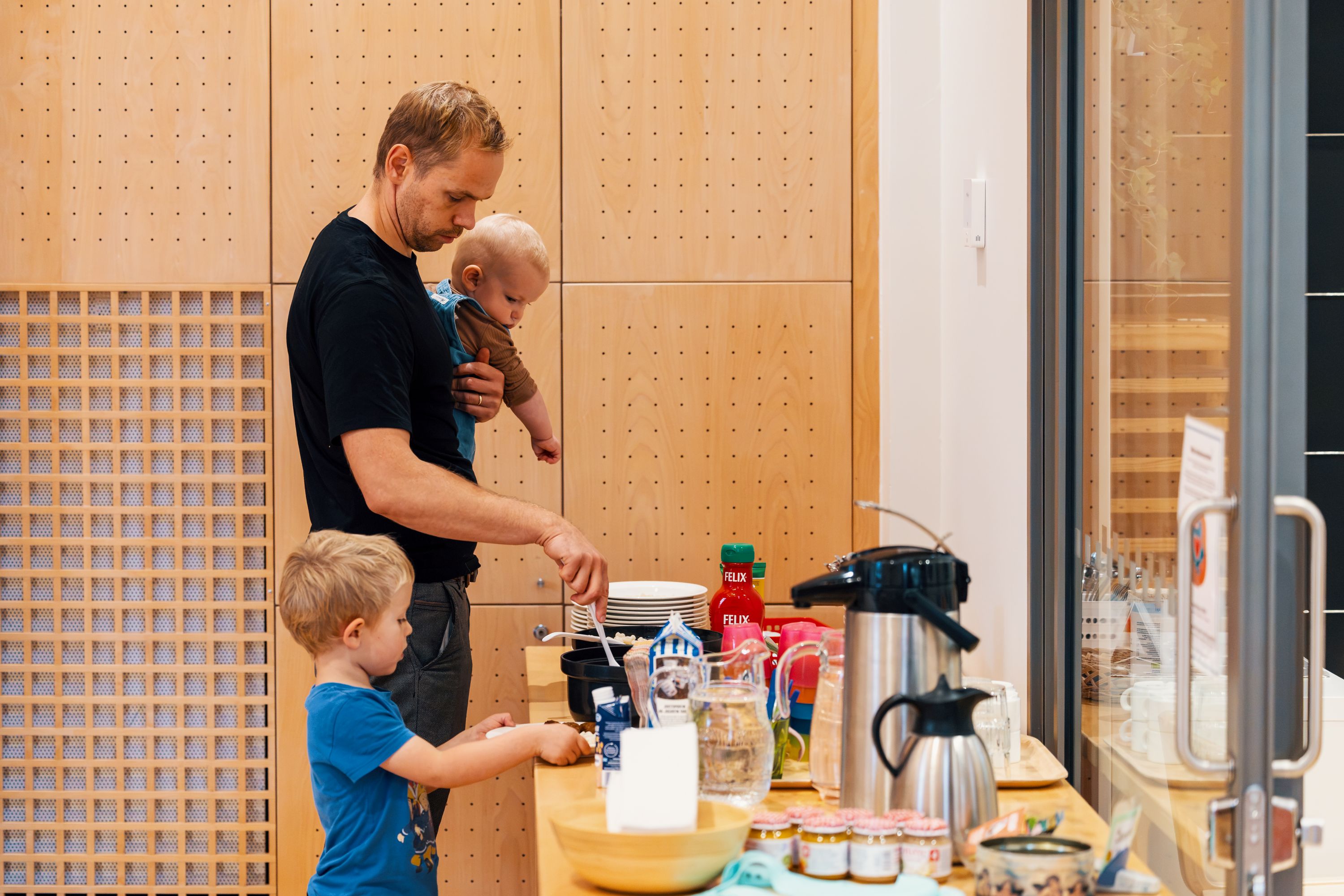 Ett barn och en vuxen tar mat från ett serveringsbord med kaffe, dryck och tilltugg. En mindre person hålls i famnen medan olika serveringskärl och matvaror står uppdukade på bordet.