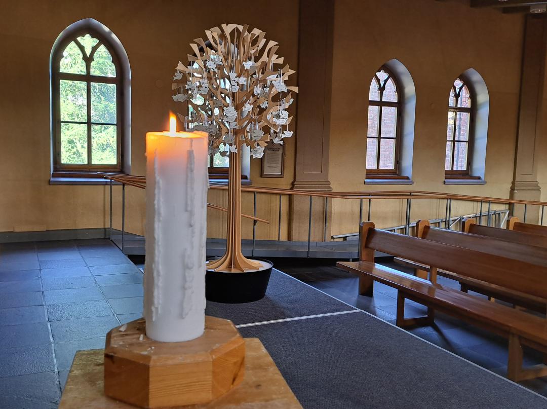 Ljus som brinner i en kyrka, i bakgrunden ett träd med dopduvor av trä i.