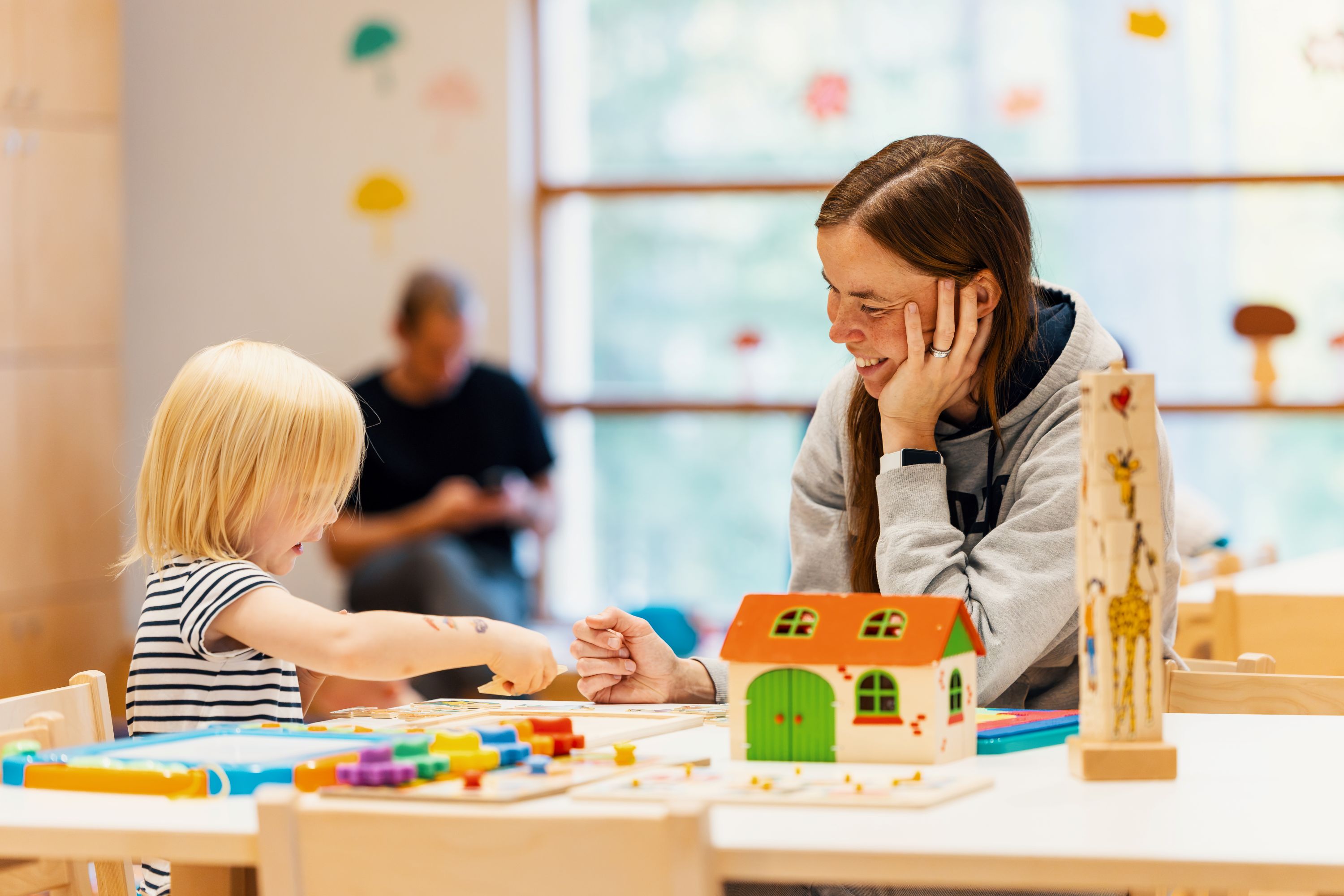 Ett barn och en vuxen sitter vid ett bord och leker med pussel och pedagogiska leksaker i ett ljust rum. På bordet står ett leksakshus och annat pysselmaterial.