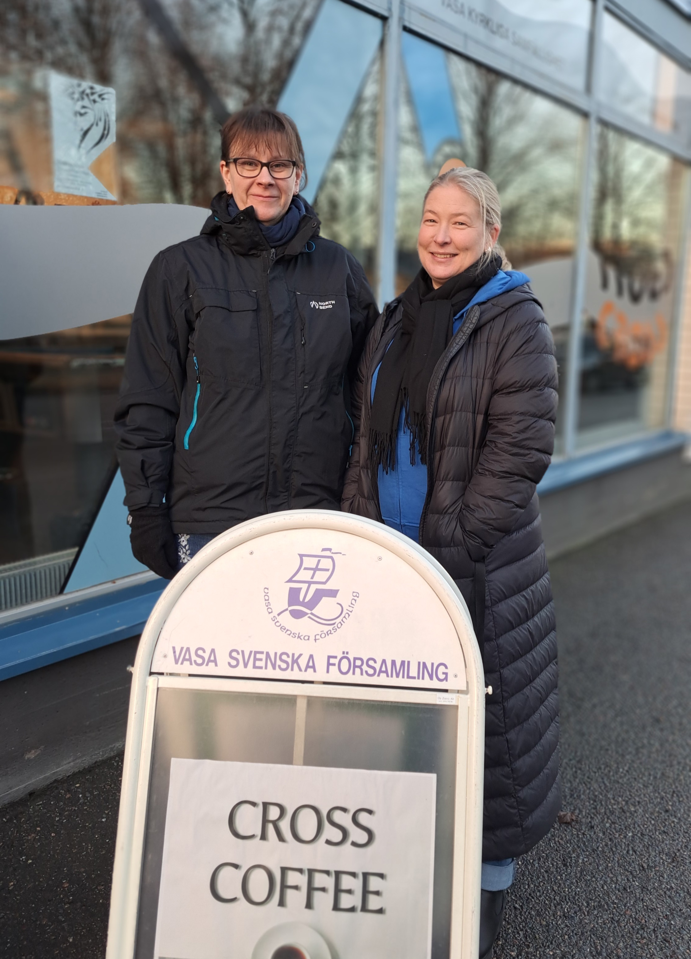 Två kvinnor står bakom en skylt med texten Cross Coffee.