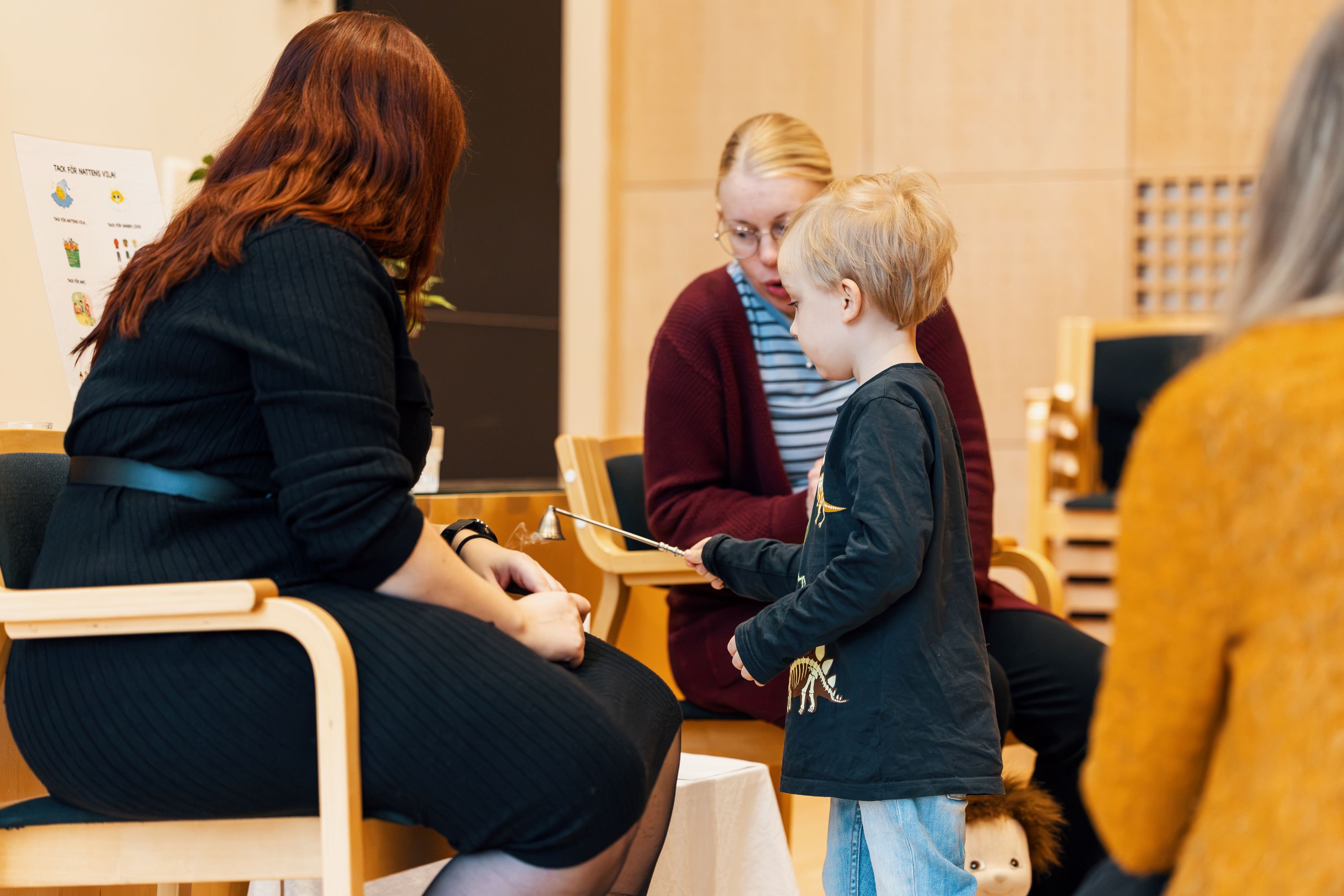 Ett barn står framför två ledare under en samling i ett församlingsutrymme och håller en ljussläckare i handen. De vuxna sitter på stolar medan barnet deltar i aktiviteten.