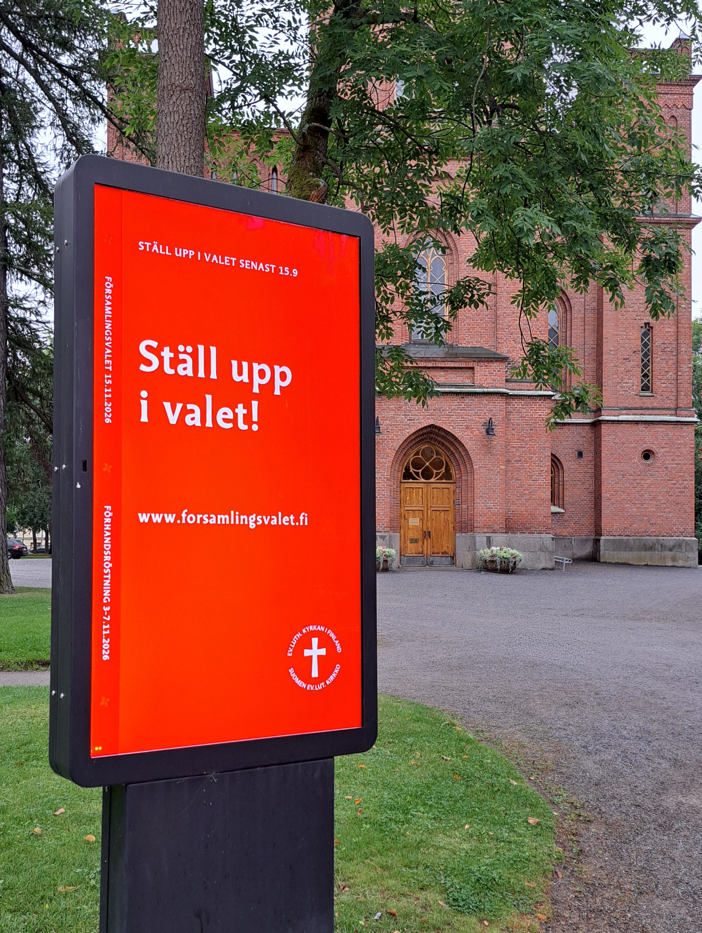 Ljustavla utanför en en röd tegelkyrka. På tavlan står: Ställ upp i valet senast 15.9!