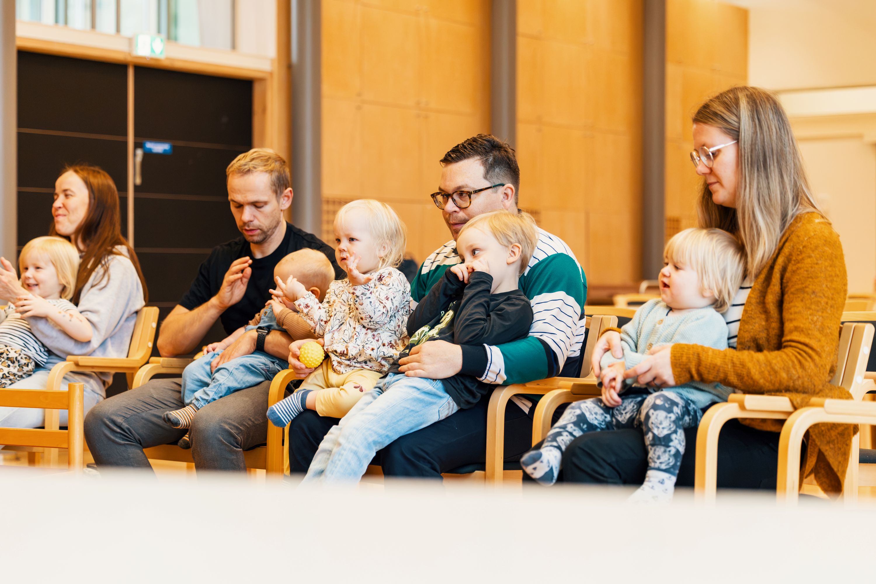 Vuxna och småbarn sitter tillsammans i en ring under en samling i ett ljust församlingsutrymme. Barnen sitter i famnen eller på egna stolar medan gruppen deltar i en gemensam aktivitet.