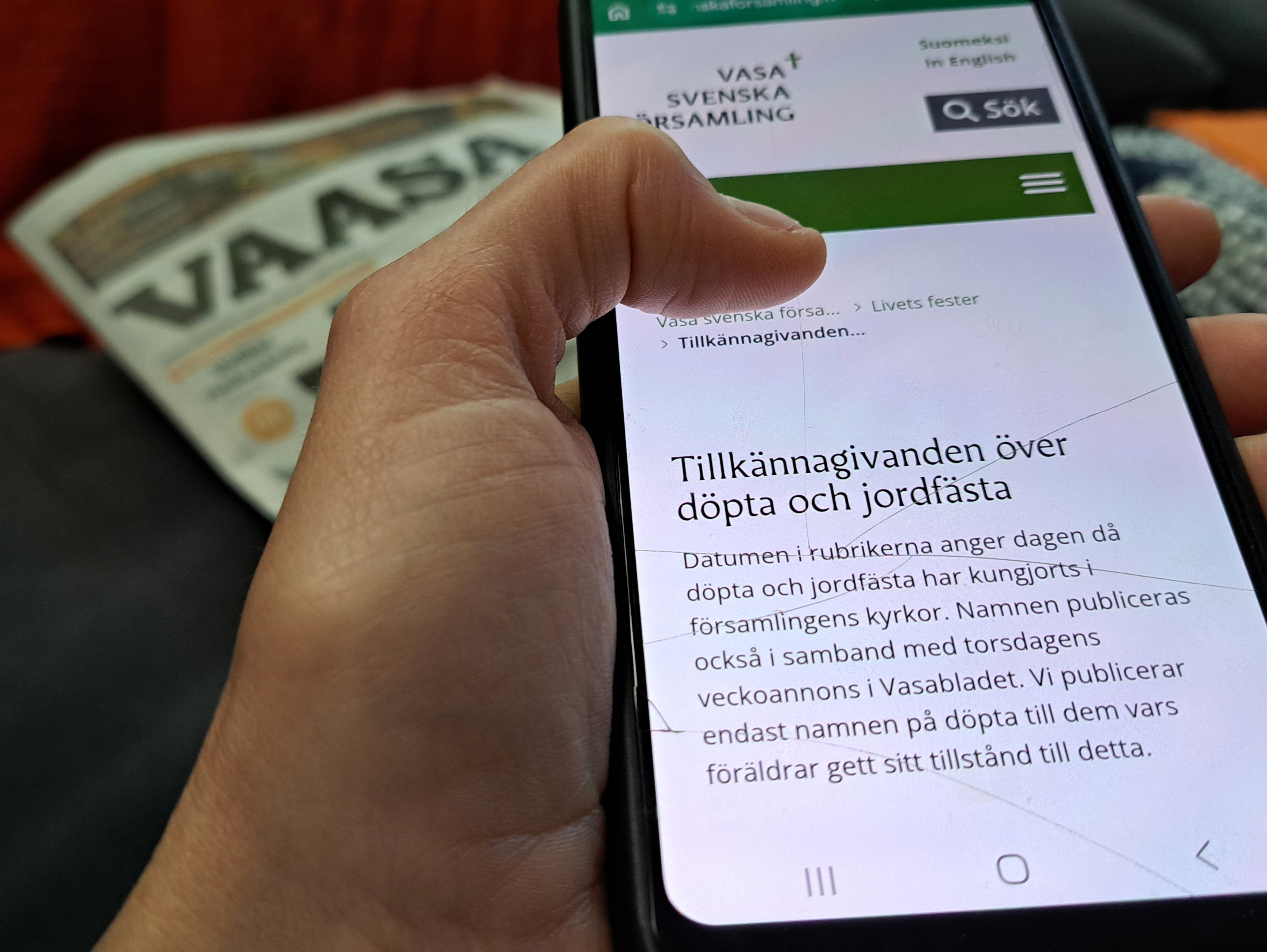 Hand håller i telefon med sida med rubriken Tillkännagivanden över döpta och döda. I bakgrunden en tidning som heter Vaasa.